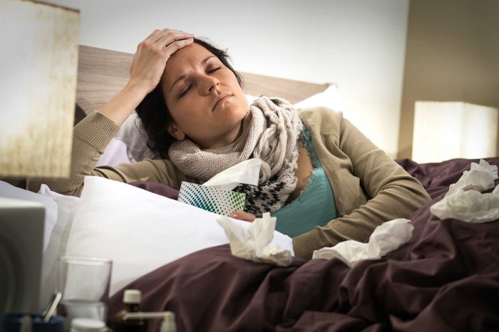 Woman lying in bed propping herself up on one elbow and holding her forehead while holding a box of tissues in the other hand.