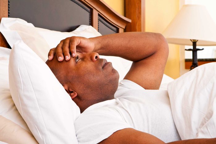 Man lying in bed resting on pillows holding his forehead with his left hand.