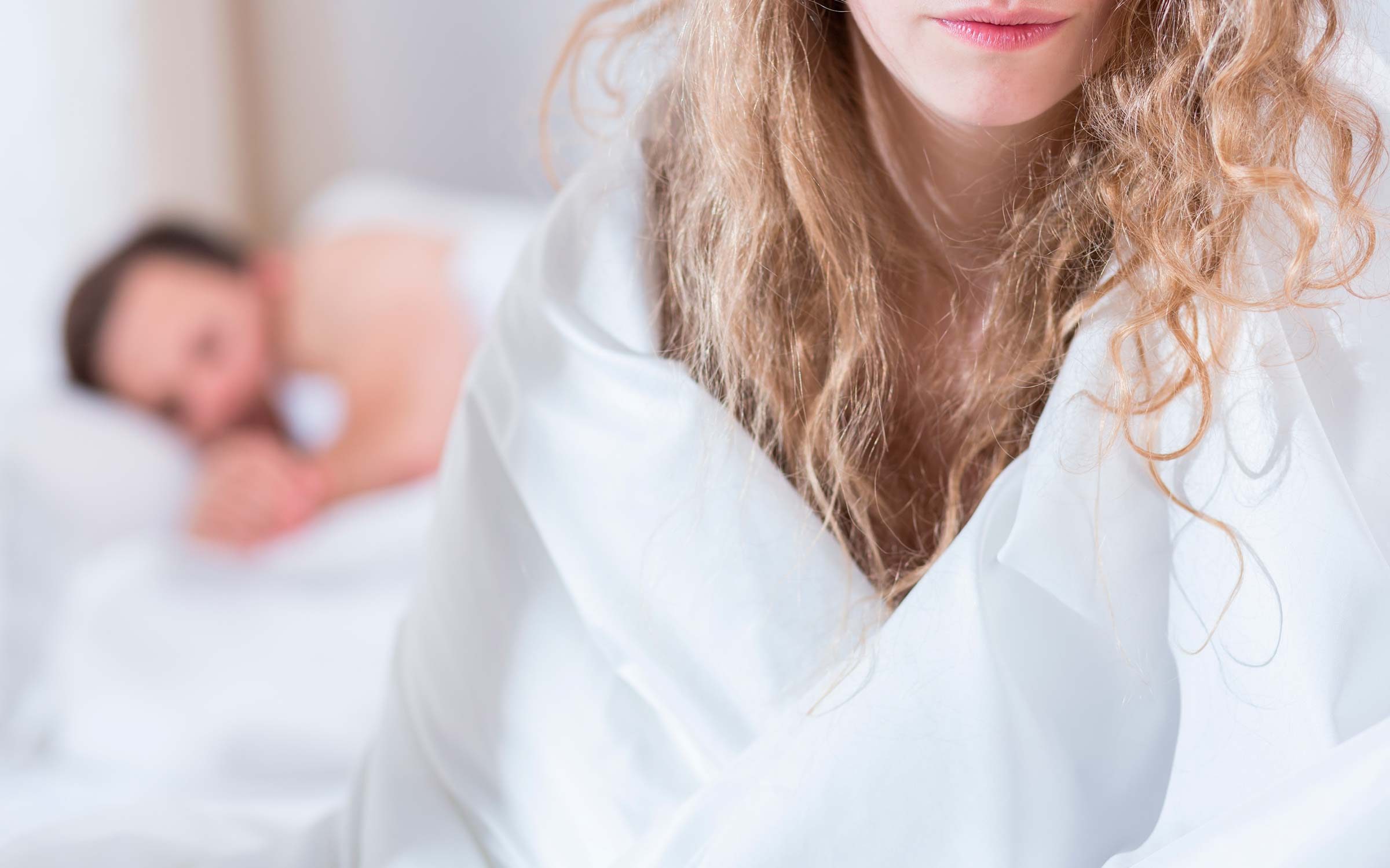 woman sitting up in bed with her back to a man blurry in the background