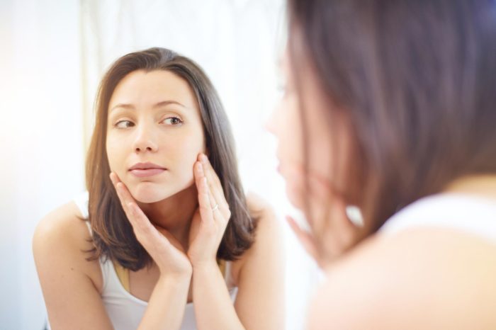 Woman in a white tank top looking at her skin in the mirror.