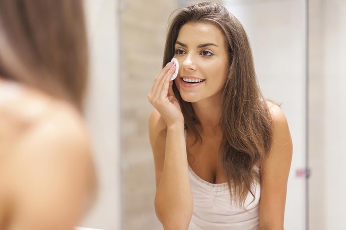 A woman applying a cleansing pad to her skin while standing in front of the mirror. 