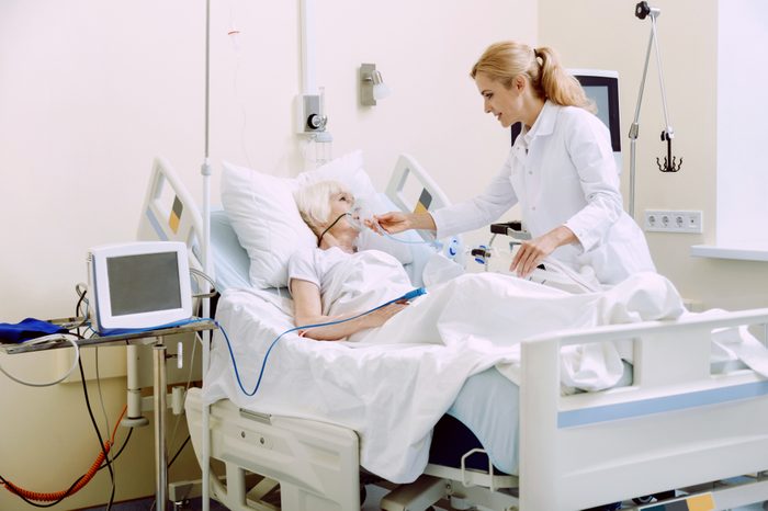 Cheerful medical worker holding an oxygen mask while adjusting it for an ill senior lady lying in a hospital bed.