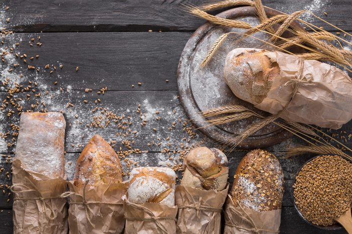 Bread background. Brown and white whole grain loaves wrapped in kraft paper composition on rustic dark wood with wheat ears scattered around. Baking and home bread making concept. Soft toning