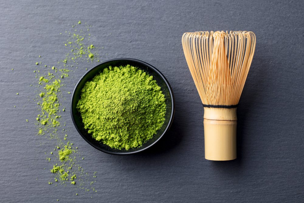 Matcha, green tea powder in black bowl with bamboo whisk on slate background. Top view.