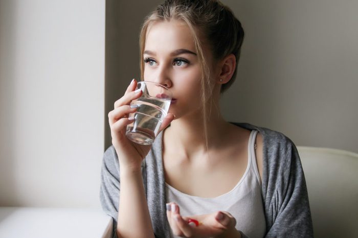 Young beautiful girl or woman take a pill or tablet with a glass of water near the window in white shirt and grey robe