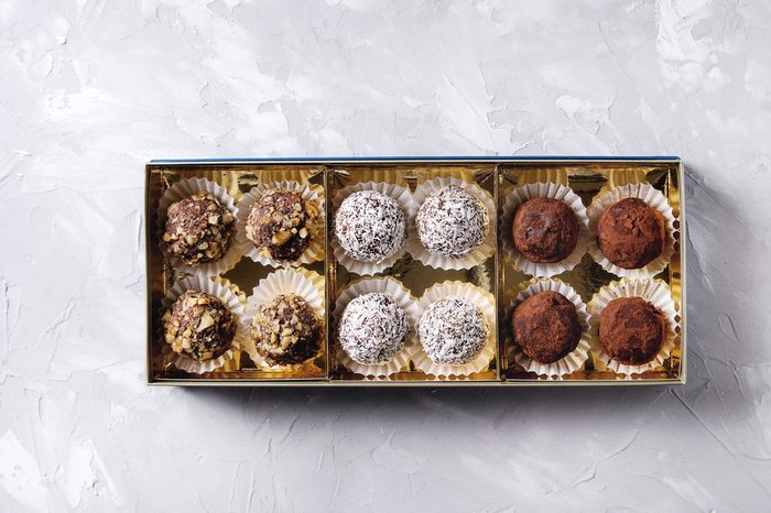 Variety of homemade dark chocolate truffles with cocoa powder, coconut, walnuts in golden gift box over gray texture background. Top view, copy space.