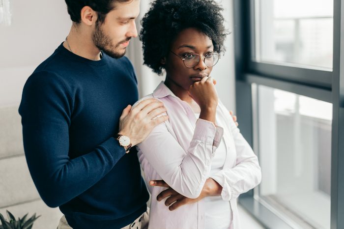 Photo of interracial couple relaxing in domestic atmosphere, have friendly talk near window in living room, dressed in casual clothes, have good relationship. Mixed race woman and man.