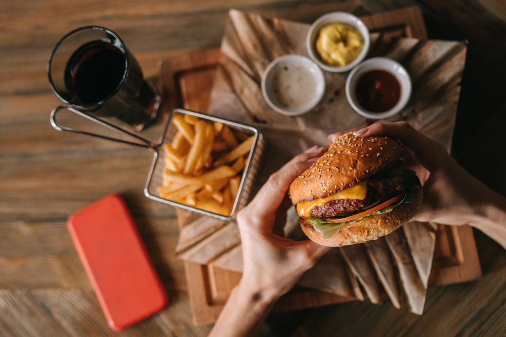 Woman holds burger with hands and fries on the background in cafe. Fresh burger cooked at barbecue in craft paper. American food. Big hamburger with meat and vegetables closeup unfocused at background