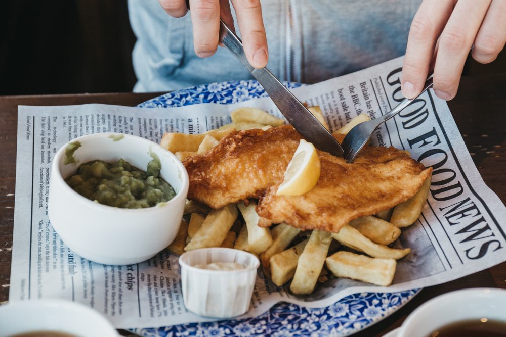 Man sitting at the wooden table, eating fish and chips, traditional English dish, tea and sauces near.