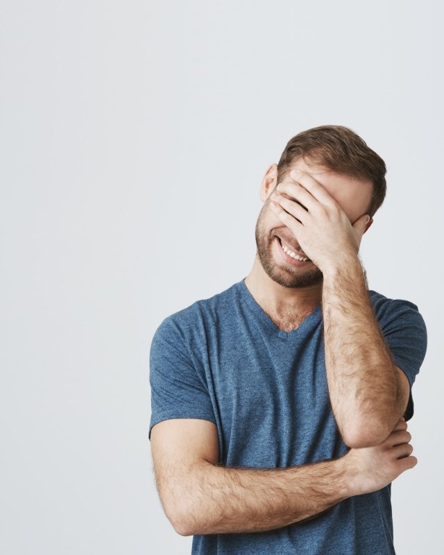 Cheerful caucasian male with stubble keeps hand on head, laughs as has fun with friends, isolated against gray background. Happy smiling guy has happy expression, hears pleasant news