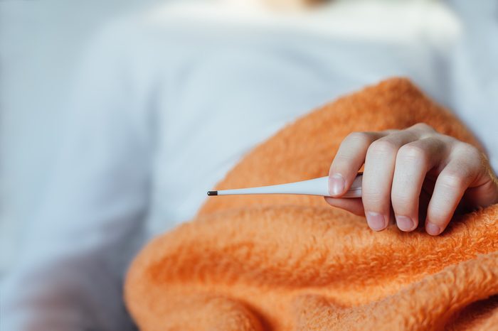 Sick man laying in bed under blanket and holding thermometer.