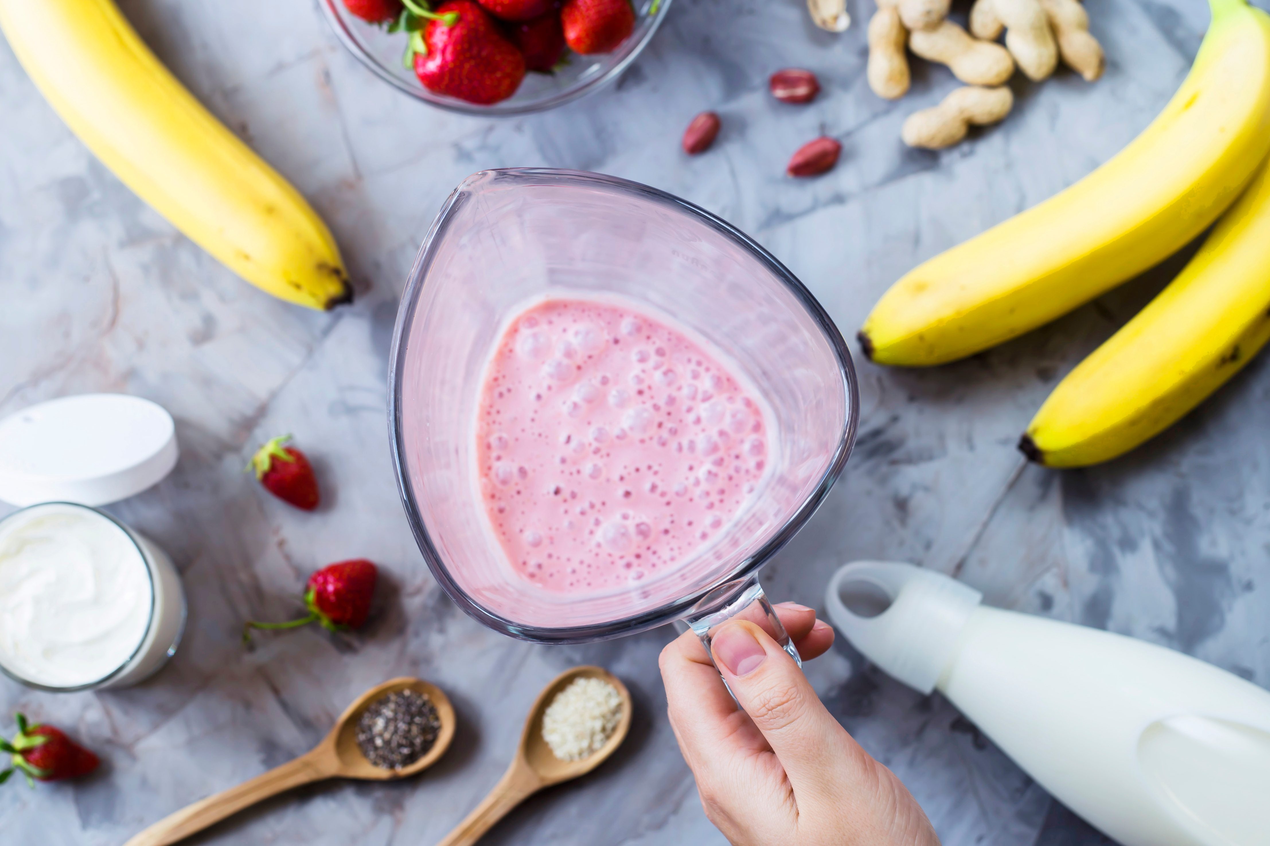 Ingredients for making strawberry banana smoothies on a gray table next to a glass bowl of blendet. Cooking a healthy breakfast concept. TOp view, flat lay