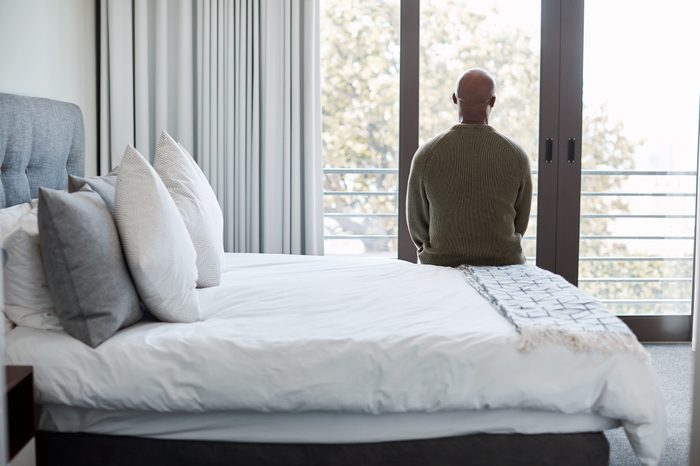 rear shot of man sitting on bed looking out window