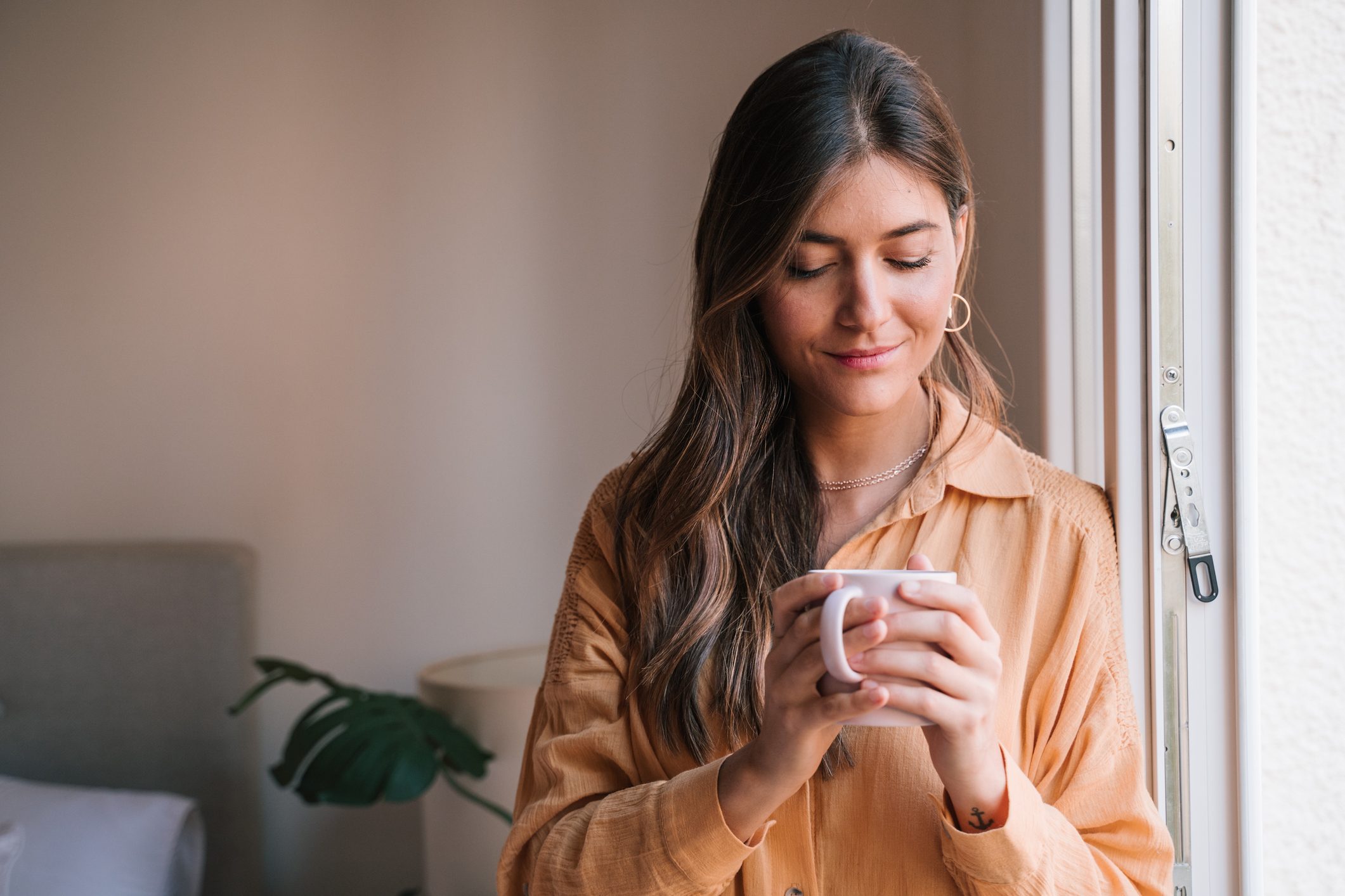 Young Woman Holding Coffee Cup By Window At Home