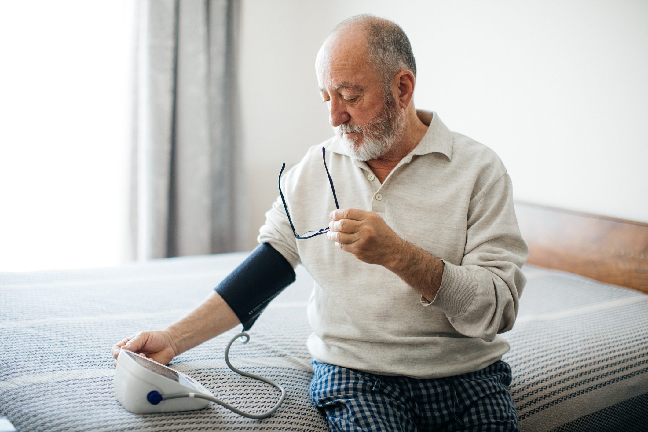 Active senior man measuring blood pressure with sphygmomanometer in bedroom