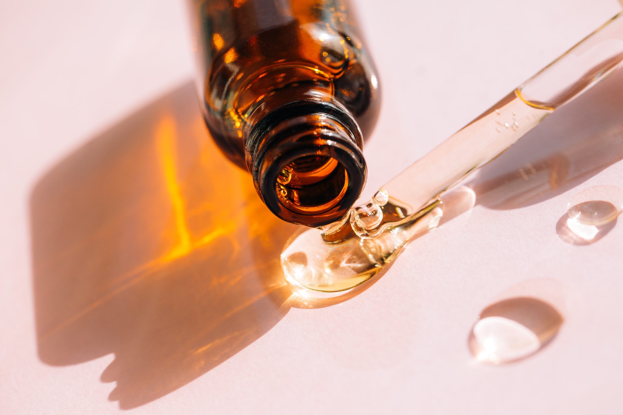 Drops of facial serum or polyglutamic acid pouring out of amber glass bottle nearby pipette on pink background. Trendy products of the year. Health and wellness concept