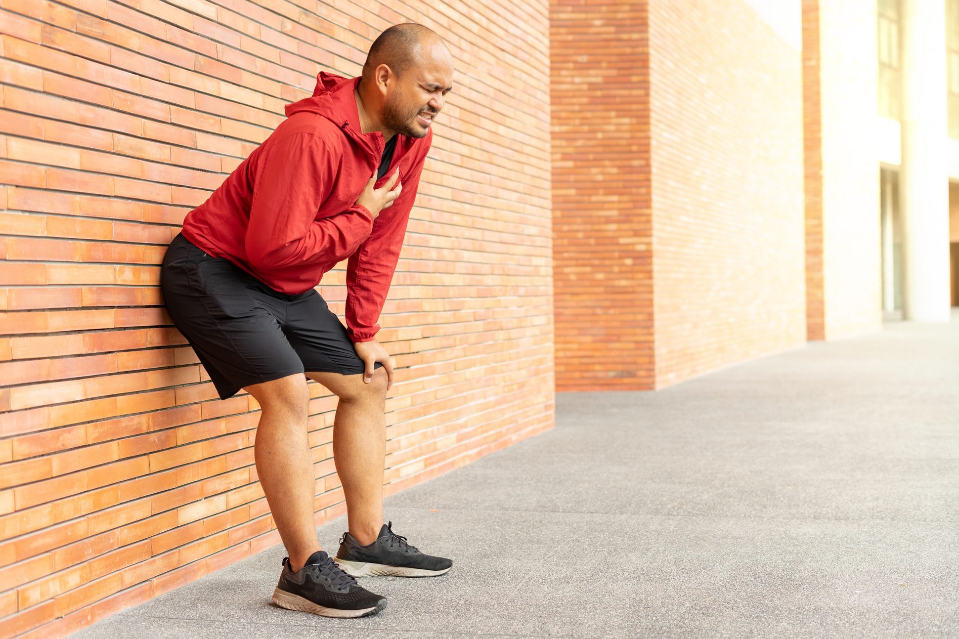 Sportsman wearing a red jacket leaning against a brick wall of clutching his chest acute pain possible heart attack. Hands male holding chest with symptom heart attack disease on city street.