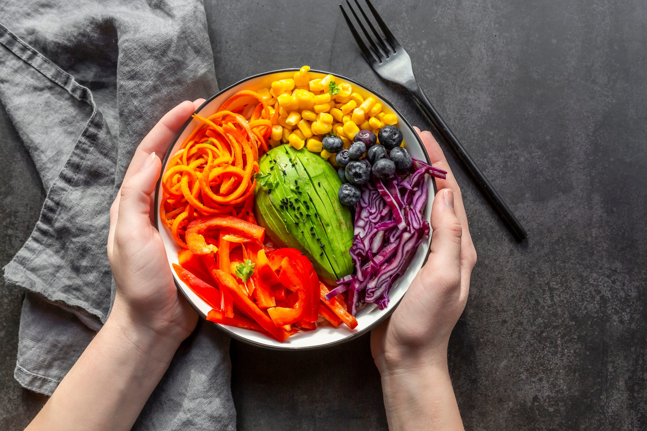 girl hands holding veggie bowl with avocado, carrots. red pepper, red cabbage, corn and blueberries