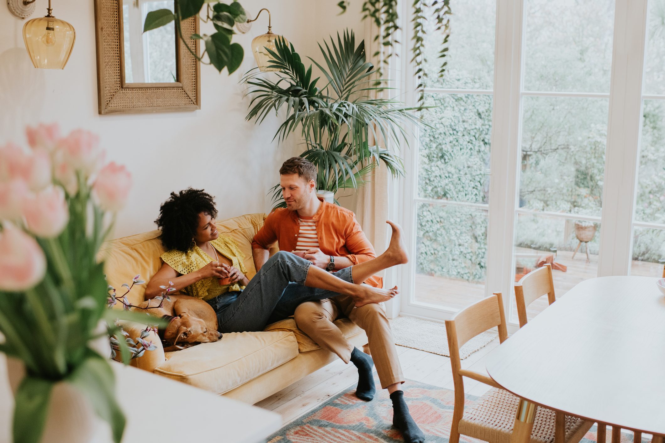 A beautiful, young, happy couple lounge together on a soft, luxurious yellow sofa in a modern living room. Their dog naps beside them.