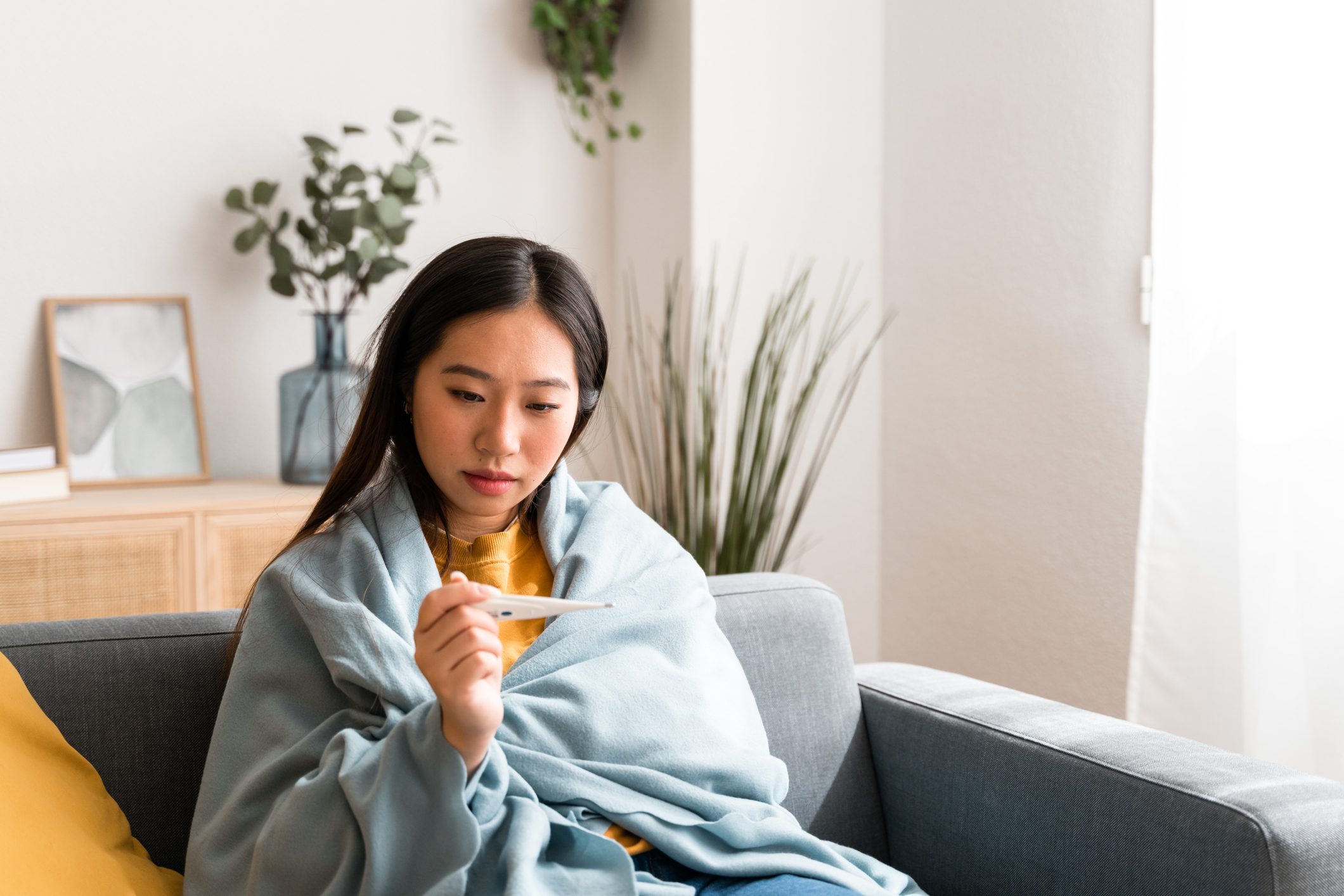 Sick woman at home covered with a blanket while using thermometer to take her temperature.